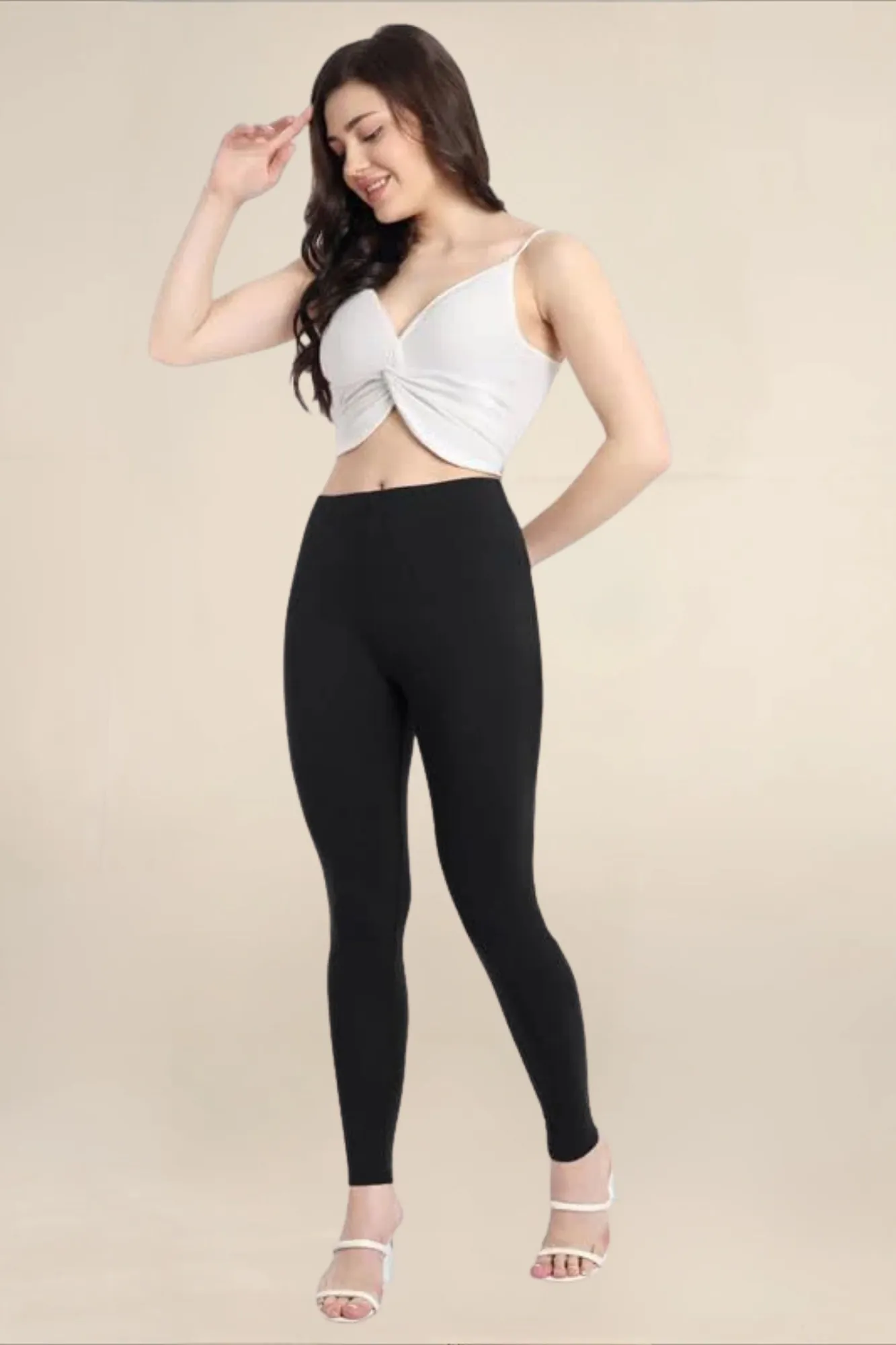 Close-up of a model posing in black high-waisted pocket leggings and white strappy block heels next to a wooden stool