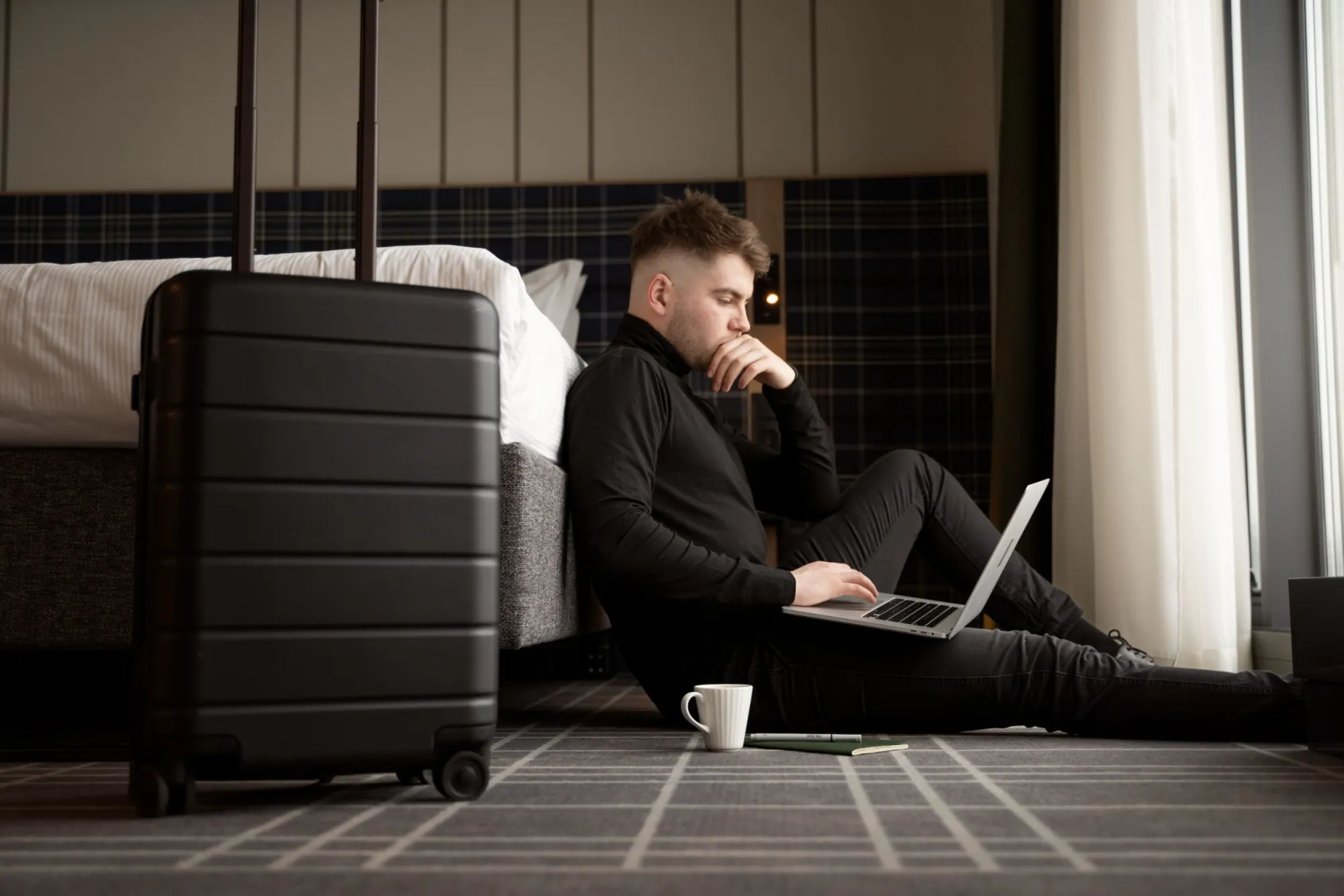 Person using a laptop on a hotel room floor beside a suitcase.