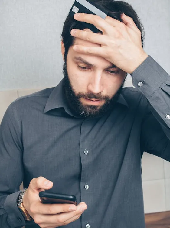 A tensed man in a black button-up shirt holding a Smartphone with his other hand on his forehead, with a credit card