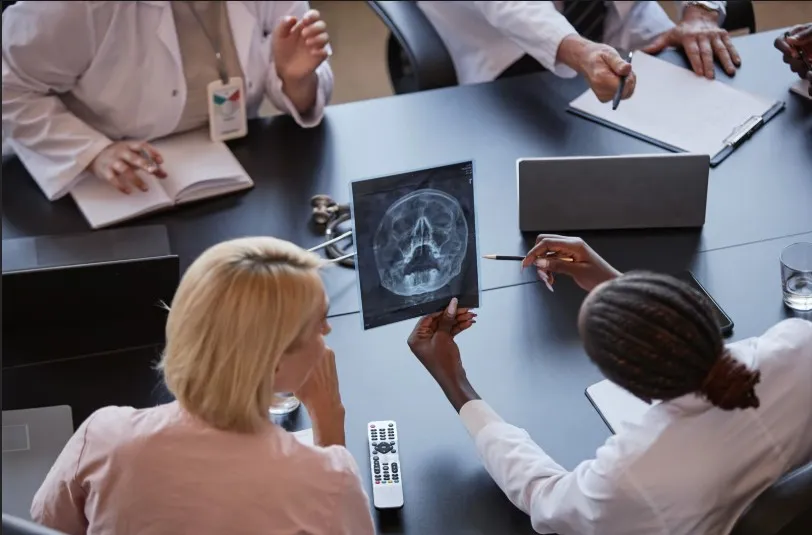 An overhead view of medical professionals in white coats sitting around a conference table discussing a skull X-ray.