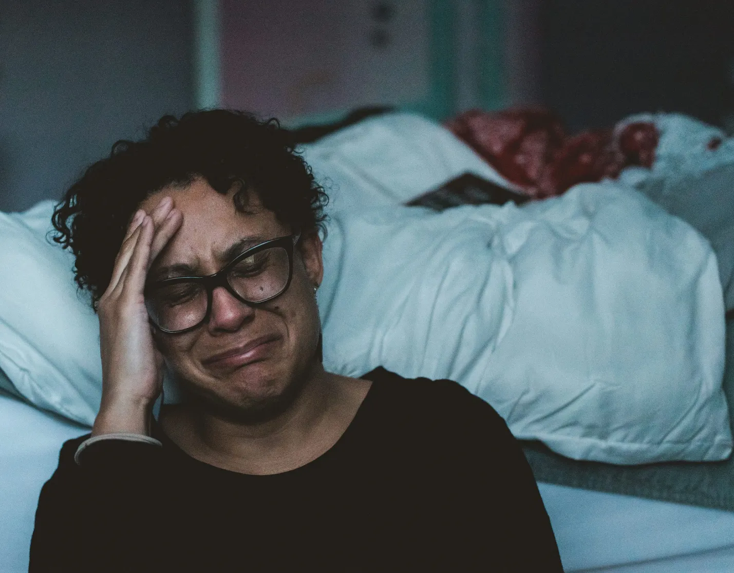 A woman sits on a bed with her hand on her head, crying, wearing glasses and a dark shirt