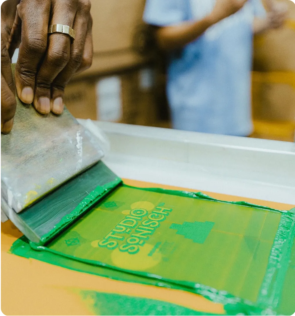 Close-up of a person using a squeegee to spread green ink across a screen-printing frame, printing the words “Studio Sonisch” onto a surface, with another person blurred in the background.