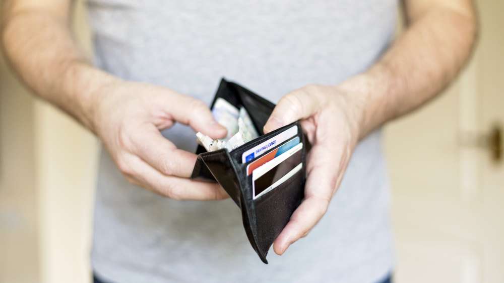 A person holding a black wallet open, showing several cards and cash inside. 