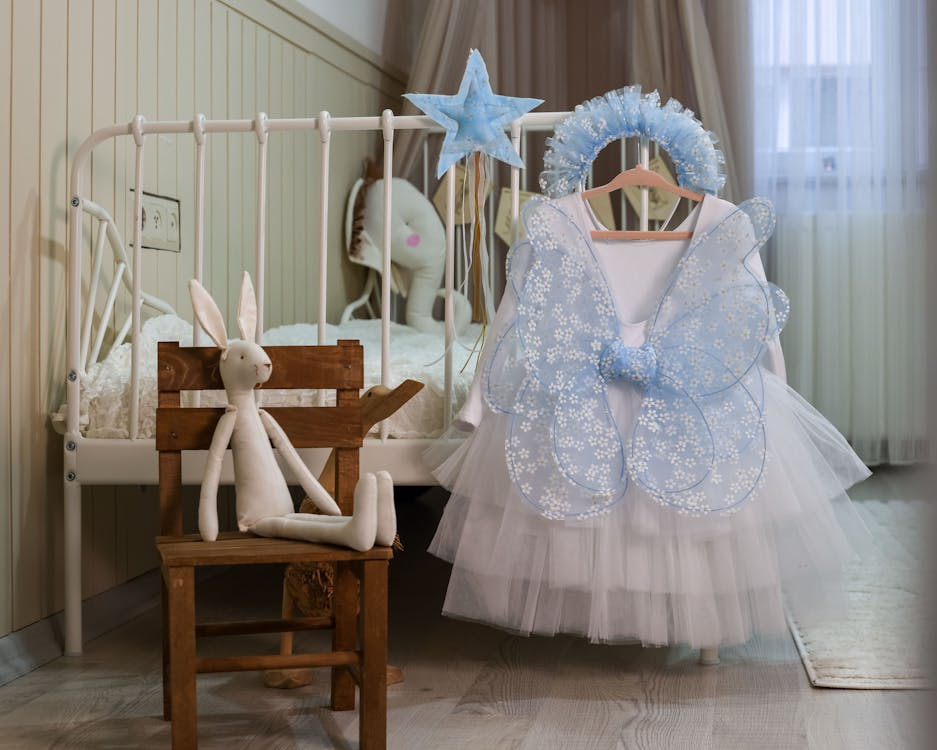A nursery scene with a white crib, soft plush toys, and a blue fairy costume hanging on display.