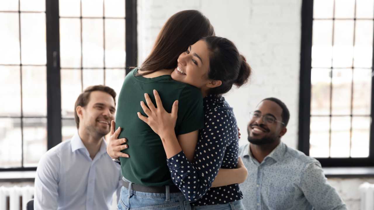 Two women hug each other happily, while two men smile in the background.