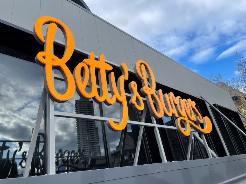 Large illuminated Betty’s Burgers building signage mounted on the exterior of a modern commercial building with reflective glass windows.