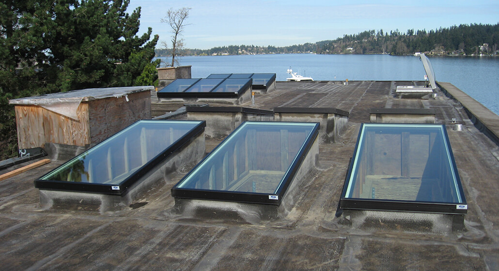 Multiple flat roof skylights with black aluminium frames installed on a flat rooftop, allowing natural light into the building, with trees and a waterfront landscape visible in the background.