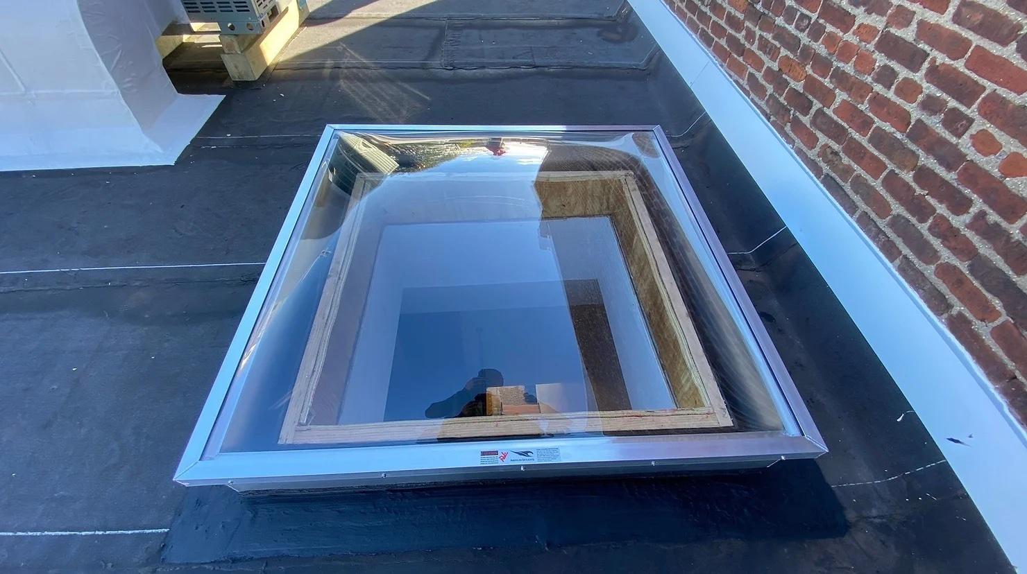 Flat roof skylight with a clear glass dome and aluminium frame installed on a residential flat roof beside a brick wall, allowing natural daylight to enter the interior space.