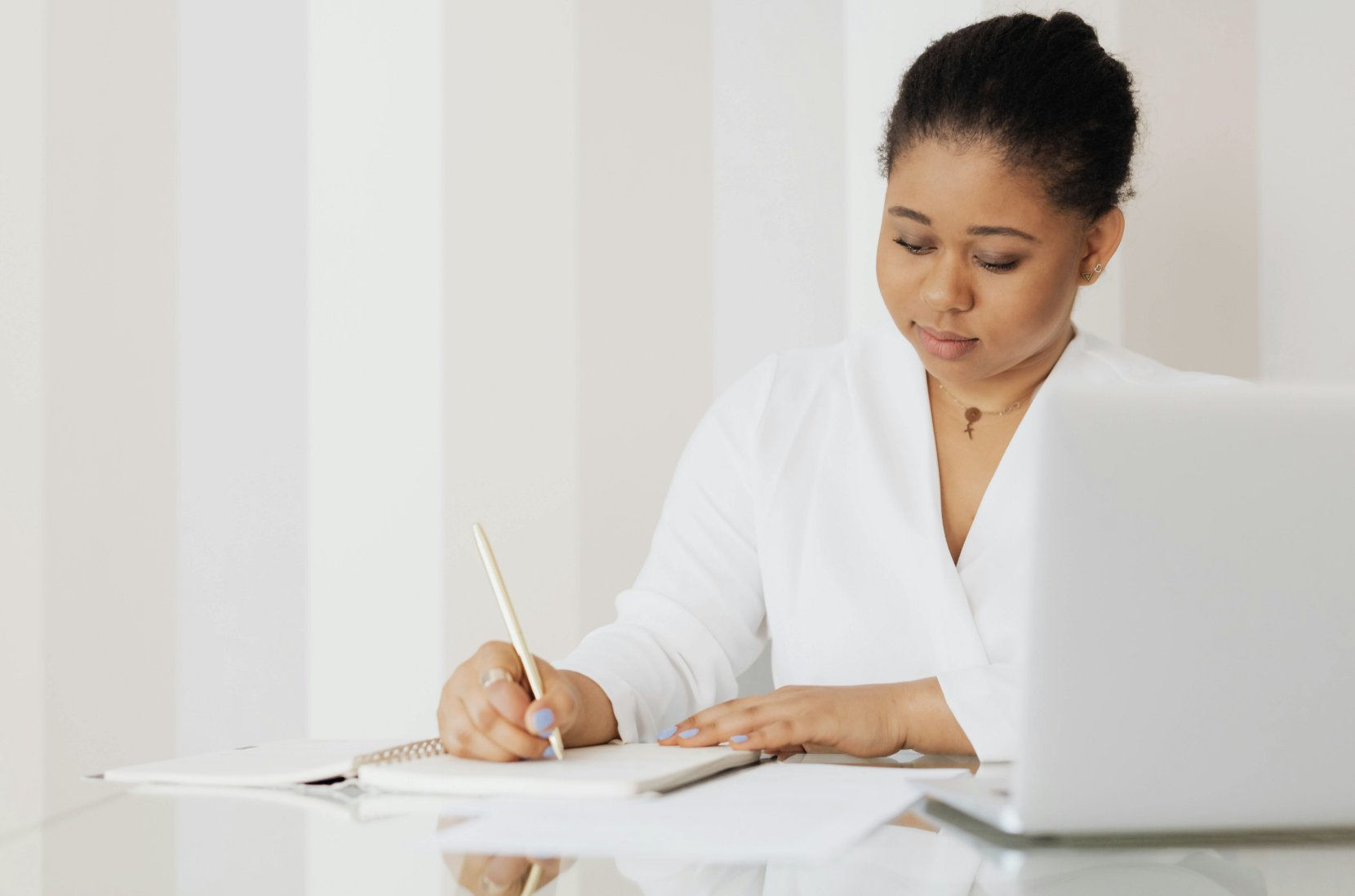 This photo shows a woman sitting at a table reading papers and writing notes in a notebook with a laptop nearby.