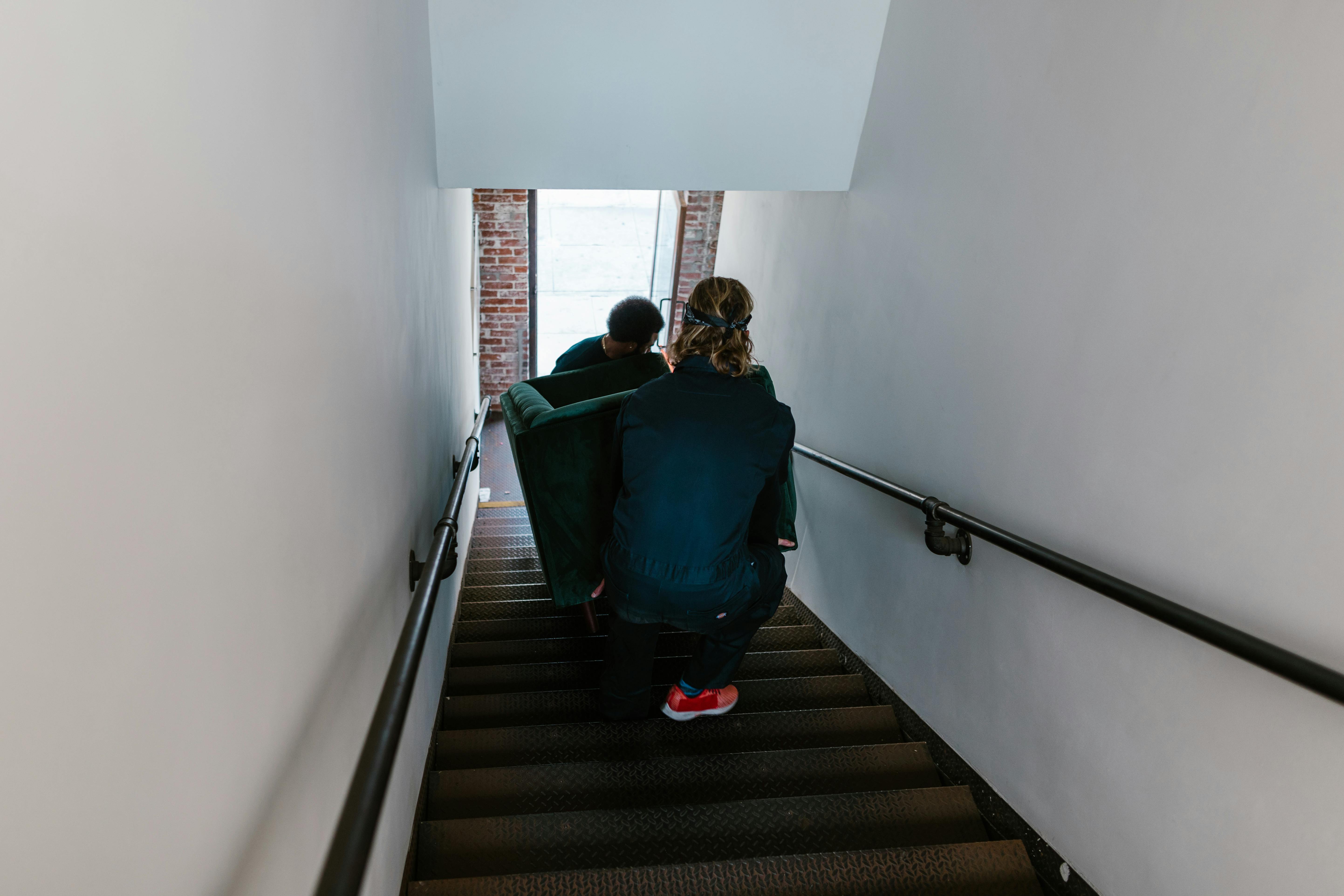 Two movers carefully carry a couch down a narrow indoor staircase during a home move.