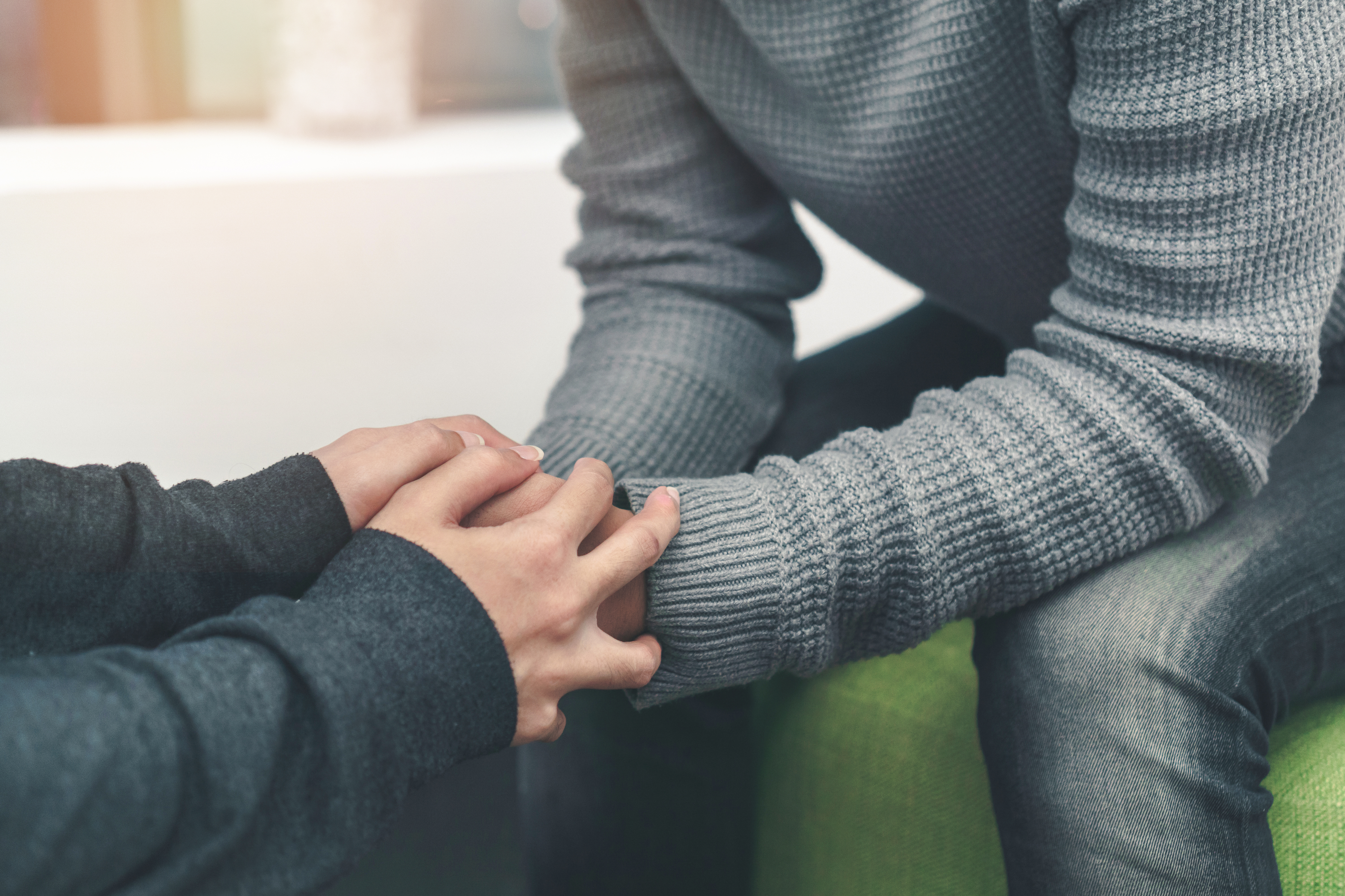 Two people holding hands, one in a gray sweater and the other in a dark sweater, sitting close together.
