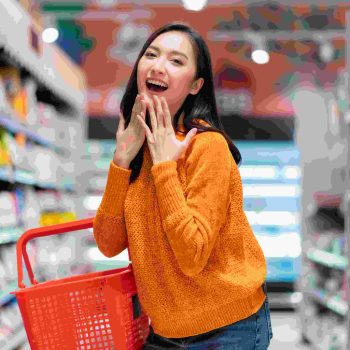 smiling-cheerful-joyful-female-woman-hand-hold-shopping-basket-hand-gesture-greeting-surprise-standing-supermarket-product-shelf-aisel-convenience-store-supermarket-department-store-mall