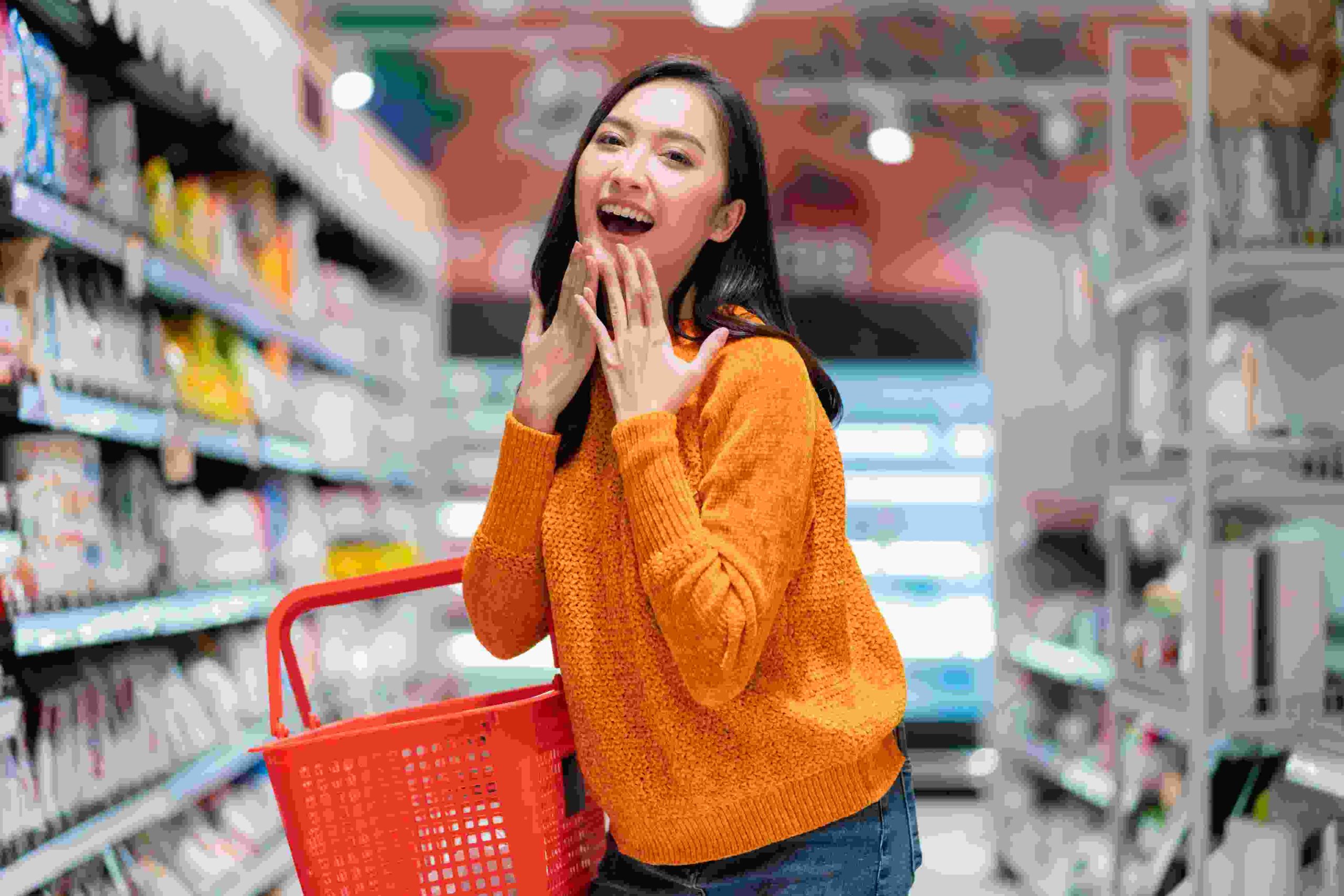 smiling-cheerful-joyful-female-woman-hand-hold-shopping-basket-hand-gesture-greeting-surprise-standing-supermarket-product-shelf-aisel-convenience-store-supermarket-department-store-mall