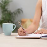 unrecognizable-woman-dress-sitting-indoors-desk-writing-journal